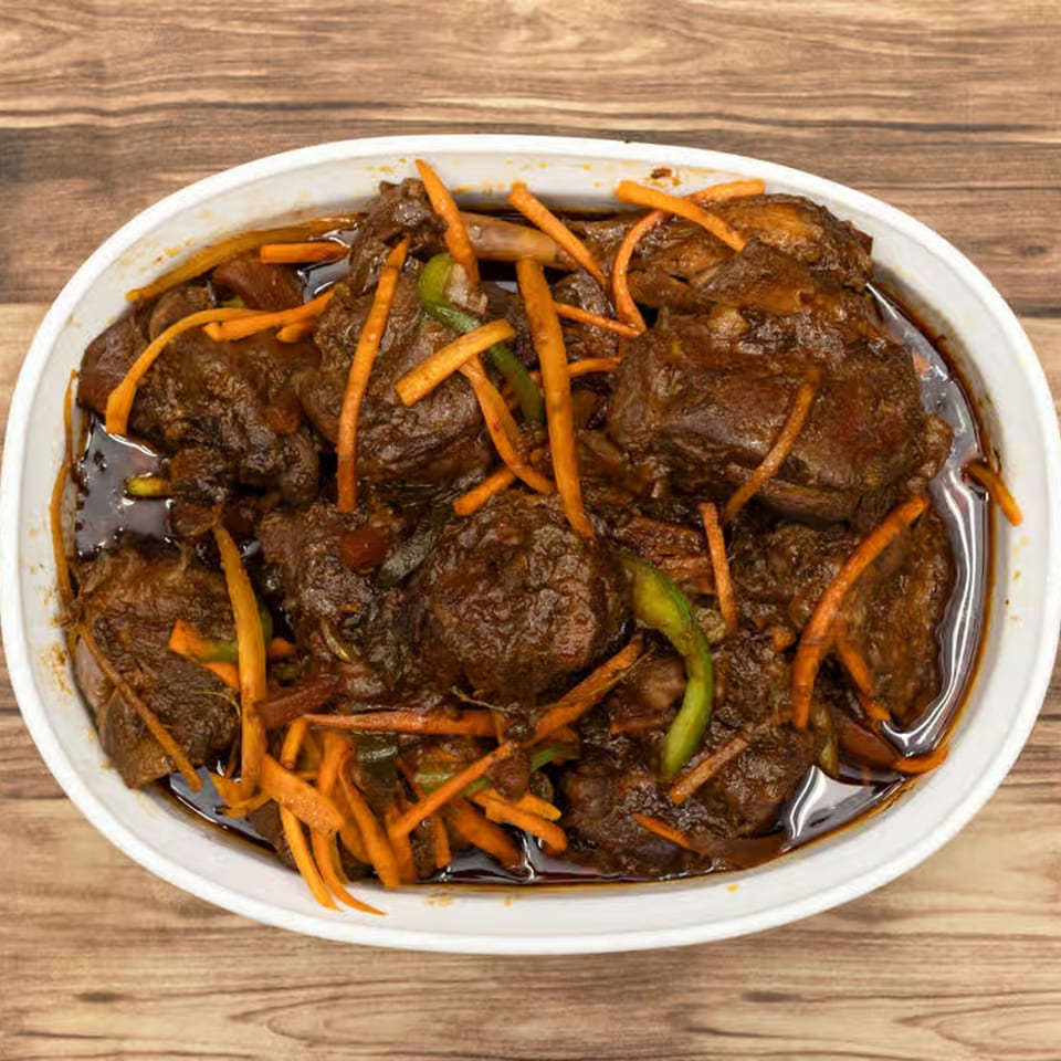 A white oval bowl filled with dark brown Jamaican stew, topped with orange carrot strips and green peppers.